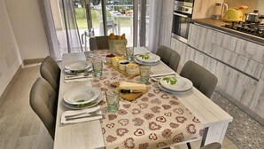 Un espace repas avec une longue table dressée pour six personnes, une cuisine équipée d’appareils modernes et une vue sur l’extérieur.