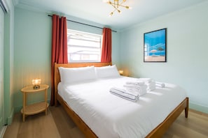 A neatly made bed with white linens and towels, a wooden headboard, a small bedside table with a lamp, and a painting on the wall. - 136 · ☀Pool✔ Kitchen✔ Beach✔ Self Check-In✔ Parking✔ (Clearwater)