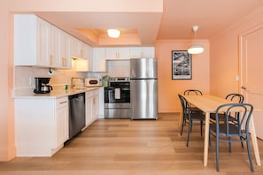 A compact kitchen with white cabinets, a stainless steel refrigerator, a microwave, an oven, and a coffee maker. There is a dining table with chairs and a pendant light above it. - 136 · ☀Pool✔ Kitchen✔ Beach✔ Self Check-In✔ Parking✔ (Clearwater)
