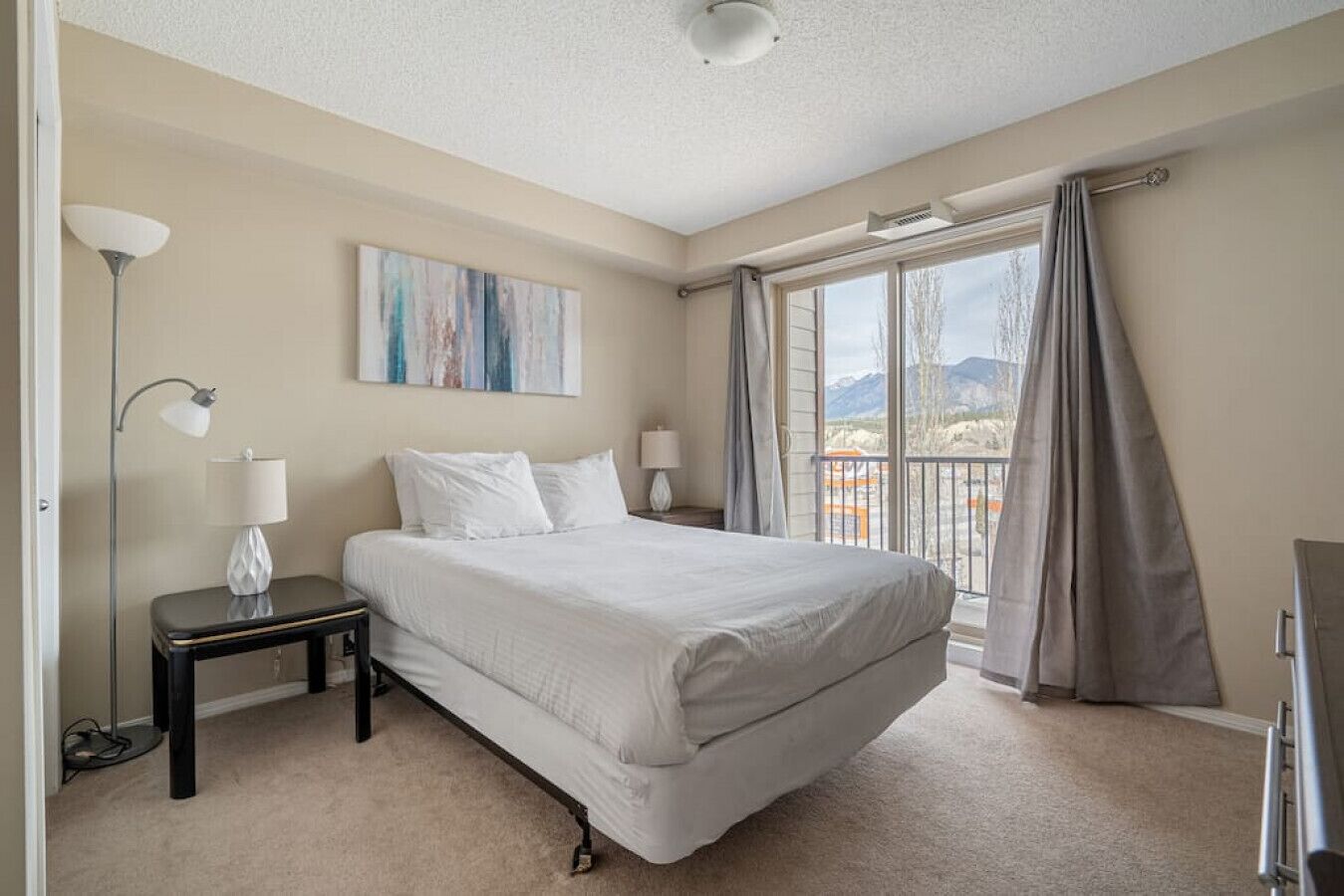 A hotel room with a large bed, a nightstand, a lamp, a painting on the wall, and a view of the mountains through the window.