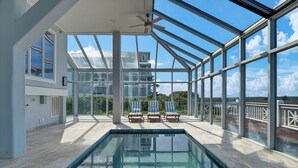Piscina interior moderna com paredes e teto de vidro, oferecendo vista para uma casa e paisagem.