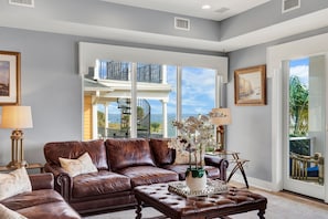 A living room with a brown leather sofa, a glass coffee table, and a view of a balcony with a railing. - Roof Top Deck w/ Ocean Views, Heated Pool Access (Tybee Island)