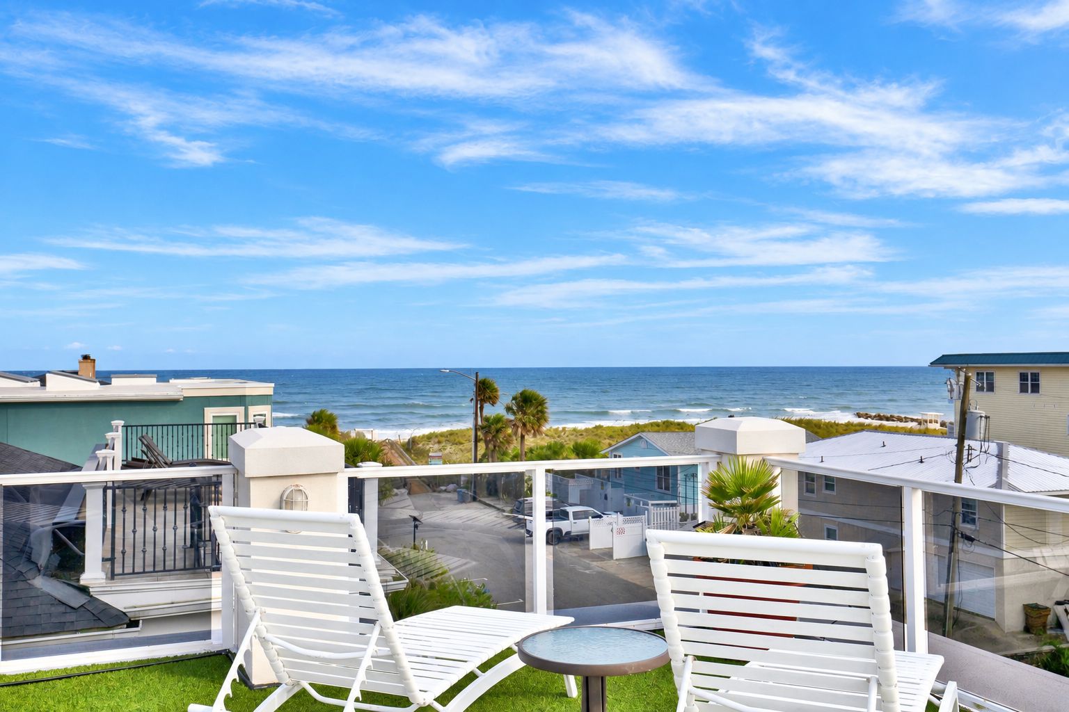 Un balcon avec des chaises blanches et une table donnant sur une plage et l’océan.