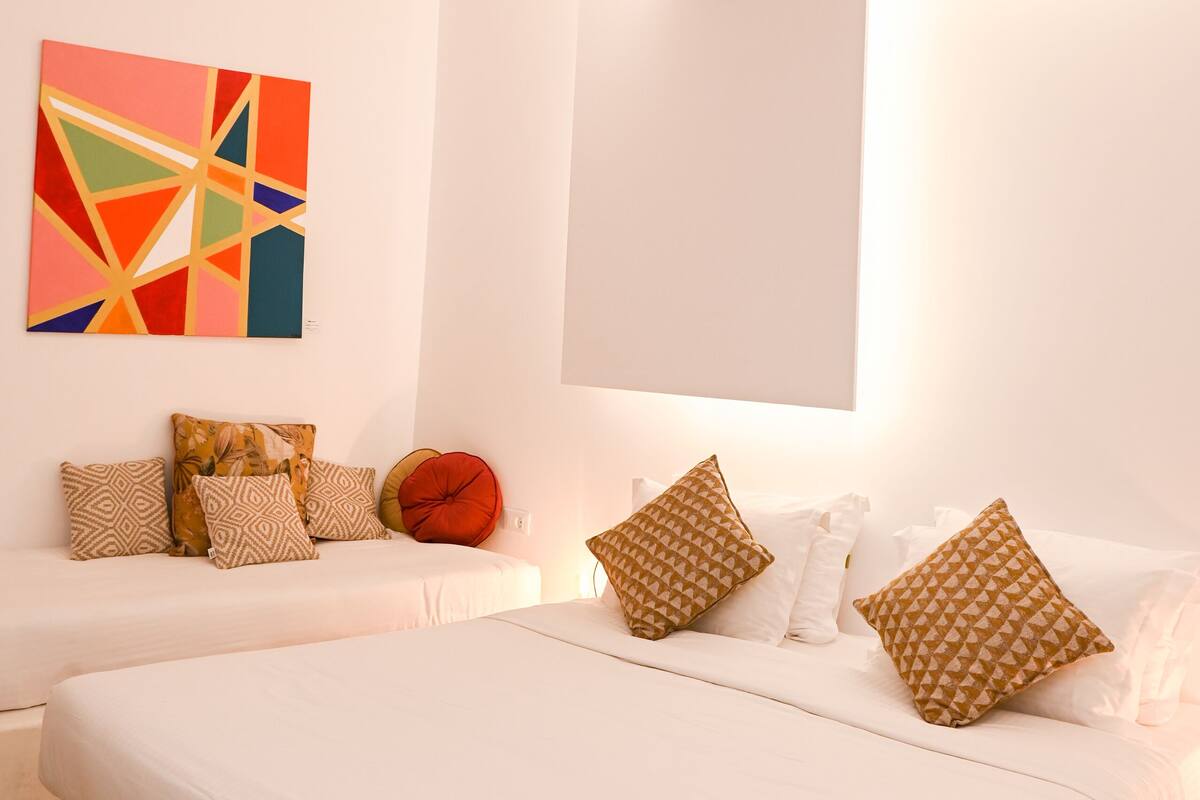 A bedroom with a large bed, two smaller beds, a colorful abstract painting, and patterned pillows.