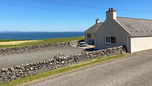 A coastal house with a stone wall, a patio with outdoor furniture, and a view of the sea and distant land.