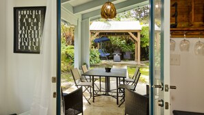 Une porte ouverte donne sur un espace extérieur abrité, aménagé avec une table et des chaises, un luminaire suspendu et un panneau avec du texte.
