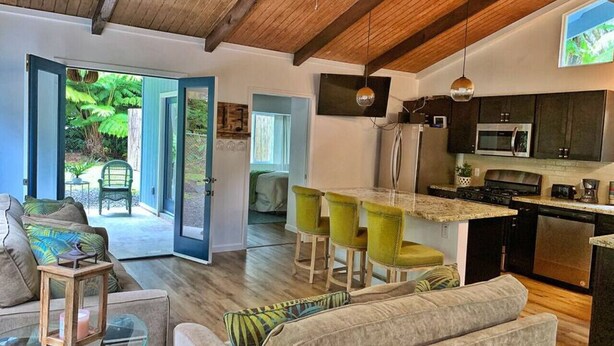 Une cuisine moderne avec un îlot central, un coin repas avec des chaises vertes et un salon accueillant avec une table basse en verre.