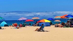 Une plage parsemée de parasols et de tentes aux couleurs vives, des gens se détendant sur le sable, et une vue dégagée sur l’océan et les montagnes lointaines.