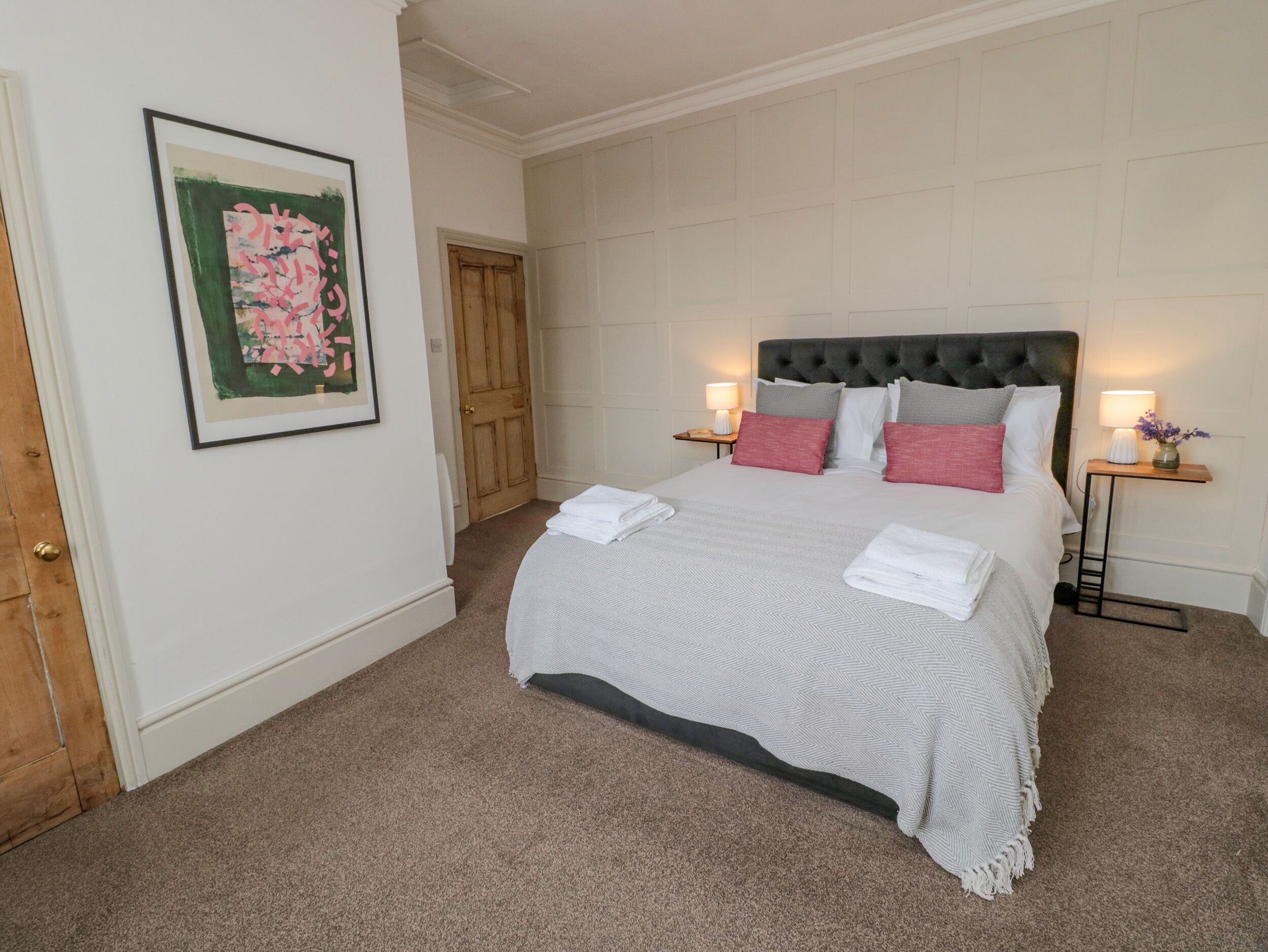 A neatly arranged bedroom with a large bed, two bedside tables with lamps, a framed artwork on the wall, and a wooden door.