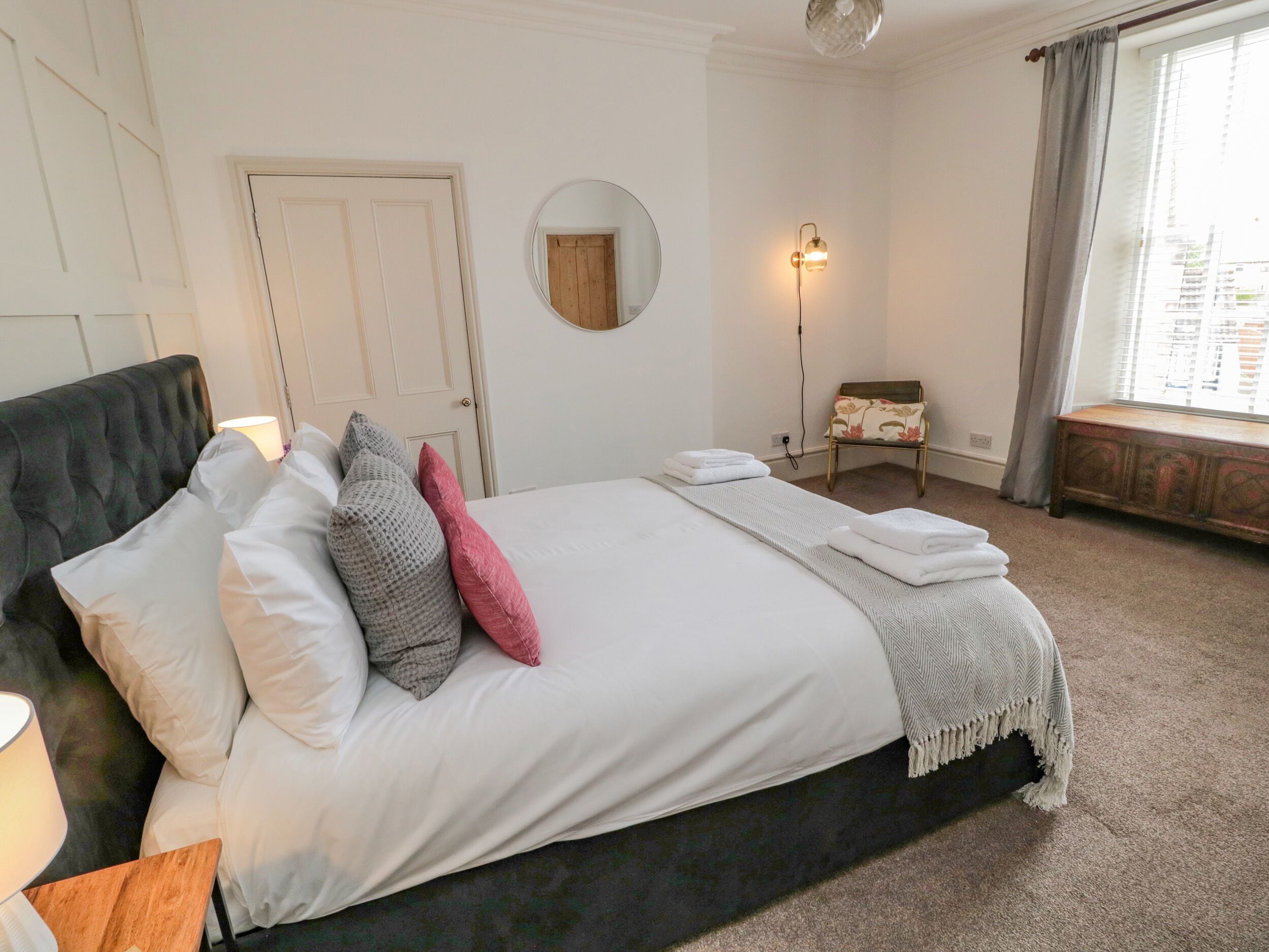 A neatly made bed with pillows, a bedside lamp, a round mirror, and a small chair with a floral pattern.