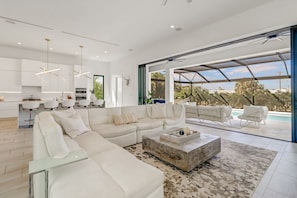 Un salon moderne avec un grand canapé d’angle, une table basse et une vue sur une piscine grâce à de grandes fenêtres.