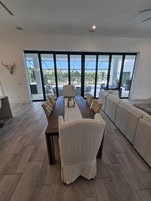 Enjoy meals with friends and family in this spacious dining area, featuring a long table and plush chairs, with a balcony offering a view of palm trees and a pool.