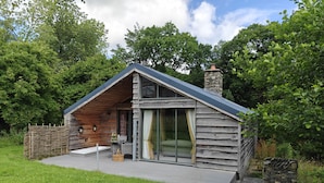 A wooden cabin with a sloped roof, a stone chimney, and a glass door leading to an outdoor seating area. - The Hare's Shack (Kendal)