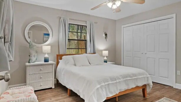 A bedroom with a bed, a ceiling fan, a window with curtains, a nightstand, a lamp, and a door. - Cedar Hideaway! (Crystal River)