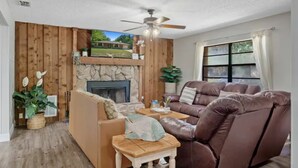 A living room with a stone fireplace, wooden panel walls, a brown leather sofa, a wooden coffee table, and a ceiling fan. - Cedar Hideaway! (Crystal River)