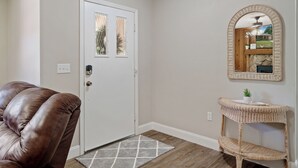 A room with a brown leather chair, a white door with glass panels, a wicker console table with a small plant, and a mirror on the wall. - Cedar Hideaway! (Crystal River)