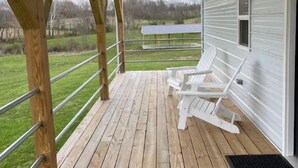 A covered porch with wooden flooring, white outdoor furniture, and a view of a green landscape. - Khyla-NEW tiny home with shared fishing pond (Athens)