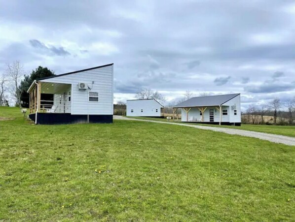A row of white single-story houses with attached porches, set in a grassy field with a cloudy sky. - Khyla-NEW tiny home with shared fishing pond (Athens)