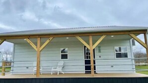 Un porche couvert avec deux chaises blanches et une petite table, une terrasse en bois et une rambarde en métal.