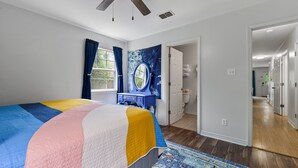 A bedroom with a bed, a ceiling fan, a window with curtains, a bathroom visible through an open door, and a hallway leading to other rooms. - Cheerful 3-bedroom private home minutes to beach (Bay St. Louis)