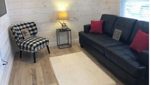 Una habitació amb una cadira amb quadres, un sofà negre amb coixins vermells, una taula petita amb una làmpada i un aire condicionat blanc al mur.