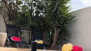 Uma garrafa de vinho e um copo sobre uma mesa de madeira, perto de uma piscina com espreguiçadeiras e almofadas tropicais.