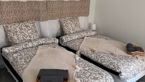 Uma cama de casal com colcha e almofadas estampadas, uma mesa de cabeceira com abajur e uma pequena cesta com toalhas.