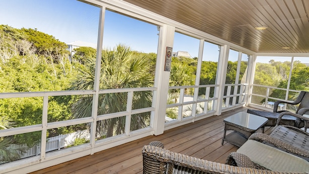 A covered outdoor area with a wooden deck, wicker furniture, and large windows offering views of greenery.