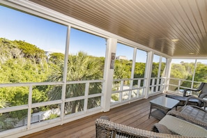 A covered outdoor area with a wooden deck, wicker furniture, and large windows offering views of greenery.