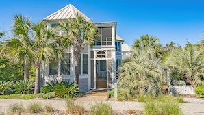 A modern two-story house with a large front porch, surrounded by palm trees and other tropical plants.