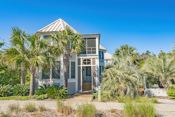 A modern two-story house with a large front porch, surrounded by palm trees and other tropical plants.