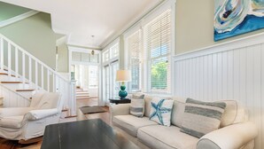 A spacious living room with a beige sofa, a black coffee table, and a white armchair. A staircase with white railings leads to an upper level.