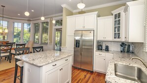 A modern kitchen with white cabinetry, a stainless steel refrigerator, granite countertops, and a dining area with wooden flooring and a table.