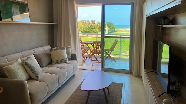 Uma sala de estar moderna com sofá, mesa de centro e televisão. Há uma varanda com vista para o oceano e vegetação.