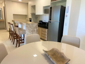 Cozinha moderna com armários brancos, geladeira de aço inoxidável e bancada de mármore. Quatro cadeiras de madeira estão dispostas ao redor de uma mesa branca.
