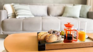 Uma bandeja de café da manhã com doces, uma cafeteira e um copo de suco de laranja sobre uma mesa de madeira em uma sala de estar com um sofá cinza.