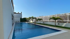 Piscina retangular com borda branca, rodeada por um muro baixo branco e uma cerca de metal.