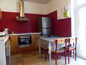 A compact kitchen with a red tiled backsplash, a stainless steel range hood, a black refrigerator, wooden cabinets, and a dining table with four chairs.