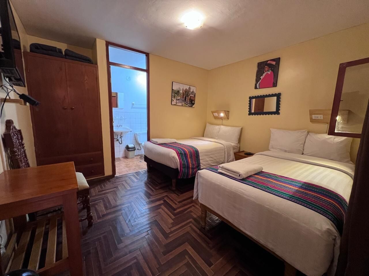 A hotel room with two beds, a wooden table, a wardrobe, and a view of the outside.