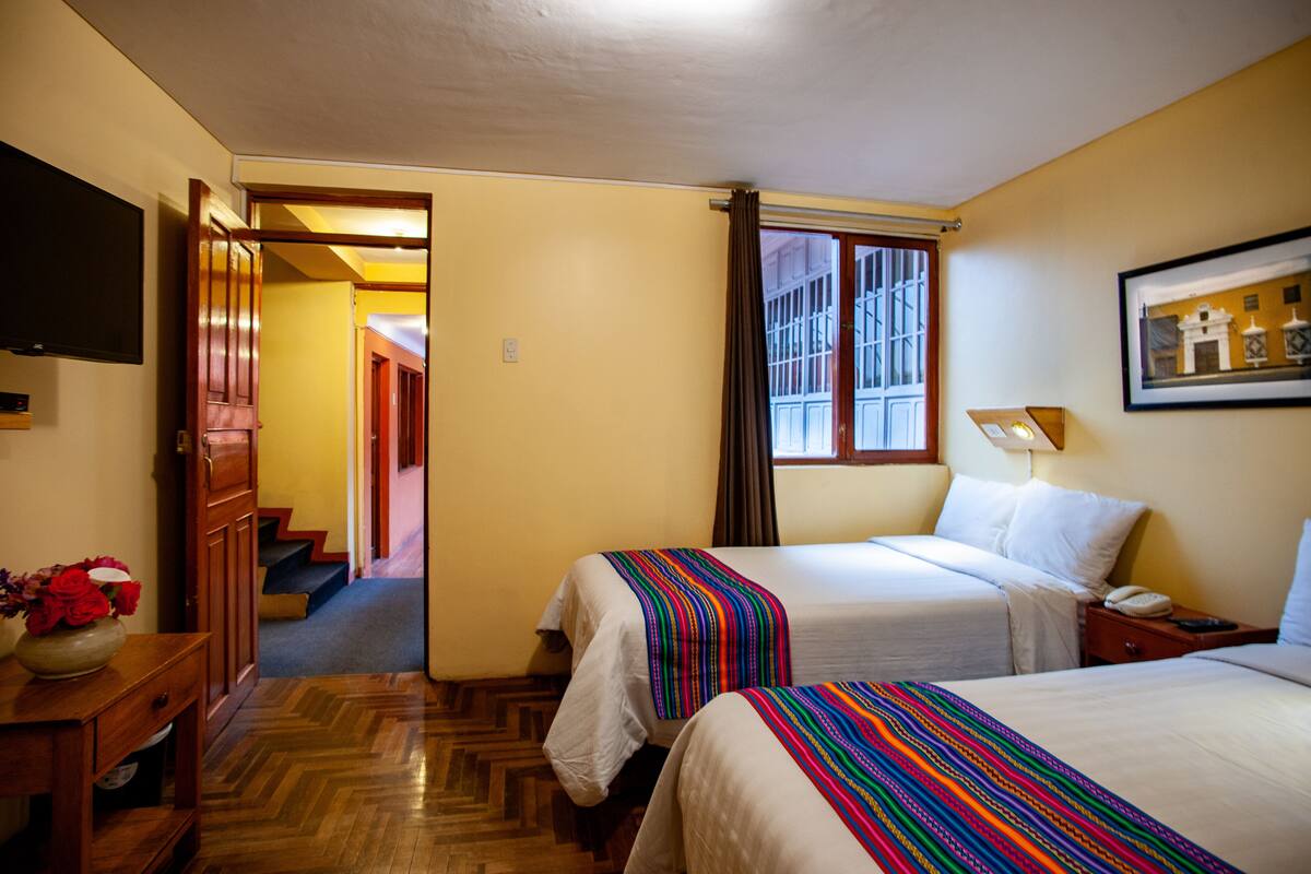 A hotel room with two beds, a TV, a desk with a vase of flowers, and a staircase leading to another room.