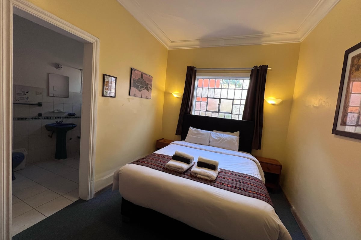 A hotel room with a bed, a window with curtains, a bedside table, and a bathroom visible through an open door.