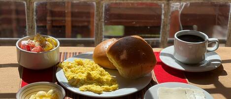 A breakfast setting with scrambled eggs, a bun, a bowl of fruit, a cup of coffee, and a plate of cheese.