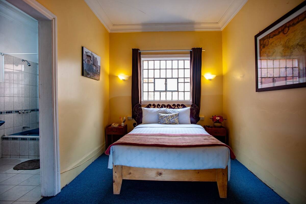 A bedroom with a wooden bed, a window with curtains, a wall-mounted picture, and a small bedside table with a lamp.