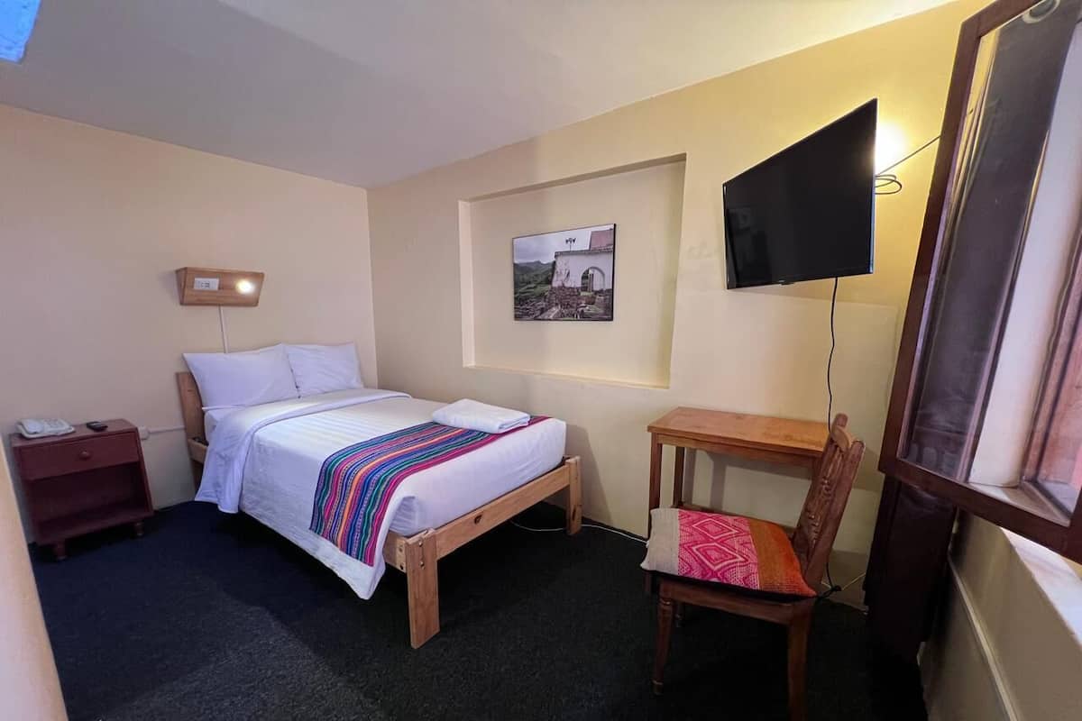 A small, neatly arranged room with a single bed, a chair, a small table, a nightstand, a wall-mounted TV, and a framed picture on the wall.