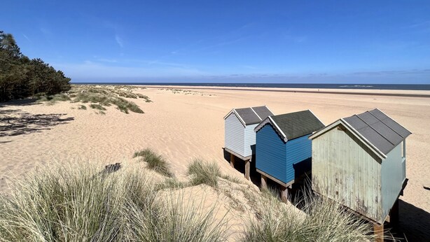 A sandy beach with three wooden beach huts and a line of trees in the background. - May Bell Cottage (King's Lynn)
