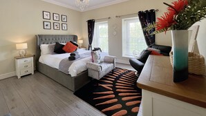 A bedroom with a large bed, a nightstand, a dresser, a chair, a chandelier, and a window with curtains. - May Bell Cottage (King's Lynn)