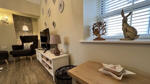 A living room with a sofa, TV, lamp, decorative plates, and a window with blinds. - May Bell Cottage (King's Lynn)