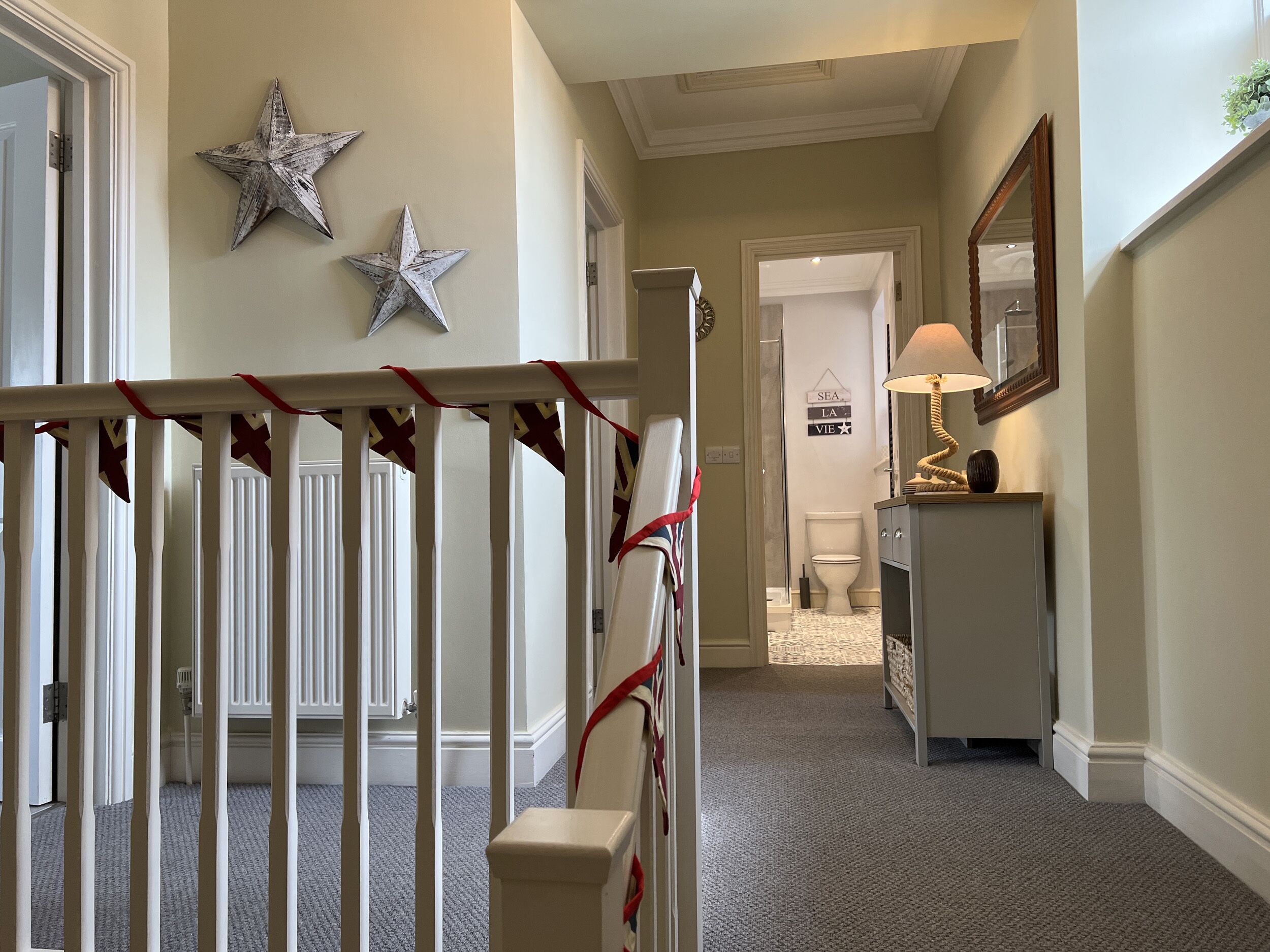 A hallway with a white railing, two star decorations, a door leading to a bathroom, and a mirror on the wall.