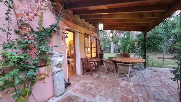 Un patio exterior con suelo de ladrillo, una mesa y sillas de madera, y una jardinera de piedra con una figura tallada.