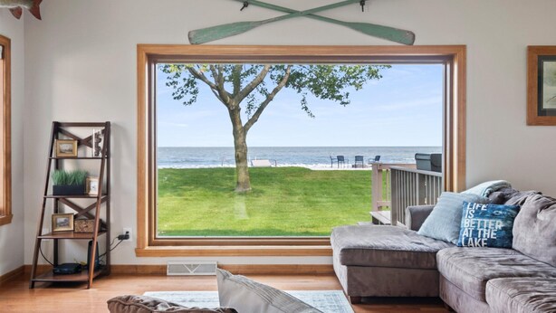 A living room with a large window overlooking a grassy area and the ocean. There is a wooden bookshelf, a gray sofa, and a framed picture on the wall.